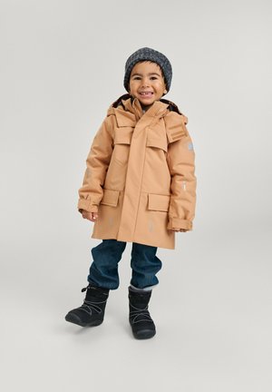 Parka impermeabile color cammello con cappuccio, diverse tasche frontali e dettagli riflettenti. Indossata su jeans blu e stivali da neve neri. Berretto di lana grigia.