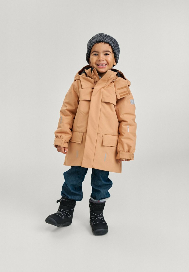 Beige wasserdichter Parka mit Kapuze, mehreren Fronttaschen und reflektierenden Akzenten. Getragen über blauen Jeans und schwarzen Schneestiefeln. Graue gestrickte Mütze.