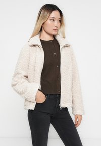 Jeune femme portant une veste en polaire beige par-dessus un pull brun à boutons et un jean noir, posant avec une main dans la poche devant un fond blanc.
