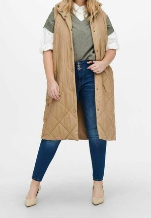 Kobieta nosząca długi beżowy pikowany bezrękawnik na szarym swetrze i białej koszuli, w połączeniu z niebieskimi obcisłymi jeansami i beżowymi szpilkami.