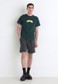 Dark green cotton t-shirt with yellow graphic design, paired with textured grey shorts. The model wears black socks and chunky black shoes.