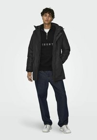 Chaqueta acolchada negra con capucha, cremallera frontal y bolsillos, llevada sobre un sudadera negra con texto blanco, combinada con jeans oscuros y zapatillas blancas.