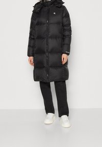 Černý péřový kabát s předním zipem, volným střihem, kapucí se stahovací šňůrkou a výraznými logy. Kombinován s černými kalhotami a bílými teniskami.