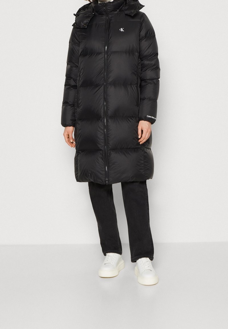 Černý péřový kabát s předním zipem, volným střihem, kapucí se stahovací šňůrkou a výraznými logy. Kombinován s černými kalhotami a bílými teniskami.