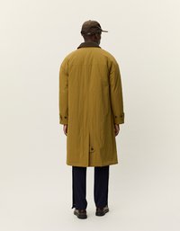 Rjav klobuk in dolg plašč gorčično rumene barve z teksturno površino, detajli z gumbi in razporki na hrbtni strani, v kombinaciji s temnimi hlačami in čevlji.