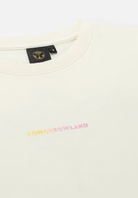 Crème-kleurig t-shirt van zachte katoen. Heeft een ronde halslijn en de tekst "TOMORROWLAND" geborduurd in gradientkleuren. Maatlabel zichtbaar.