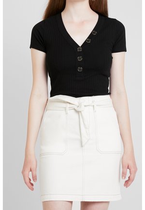 Vrouw draagt een zwart kort mouwtjes knoopjes V-hals topje dat ingestopt is in een witte hoge taille rok met een gestrikte riem in de taille.