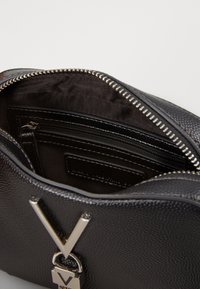 Borsa a mano in pelle nera con una finitura texture, dettaglio hardware a forma di V in argento, chiusura con zip e scomparti interni. Fodera liscia, design compatto.