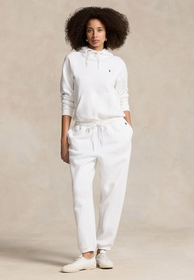 Femme aux cheveux bouclés portant un sweat à capuche blanc, un pantalon de survêtement blanc et des baskets blanches, debout devant un fond beige uni.