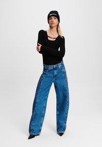 Zwarte geribbelde top met lange mouwen en een ronde halslijn, hoge taille, blauwe wijde jeans, aangevuld met een zwarte gebreide beanie met logo tekst.