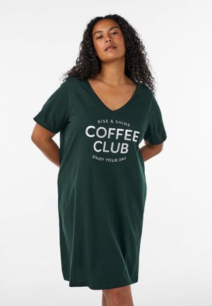Femme aux cheveux bouclés portant une robe vert foncé avec le texte "Rise & Shine Coffee Club Enjoy Your Day" sur le devant, debout les mains derrière le dos.