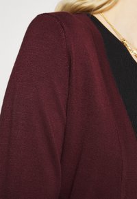 Burgundský cardigan s texturovaným pletením a dlouhými rukávy, navržený s výstřihem do V a vrstvený přes černý top.