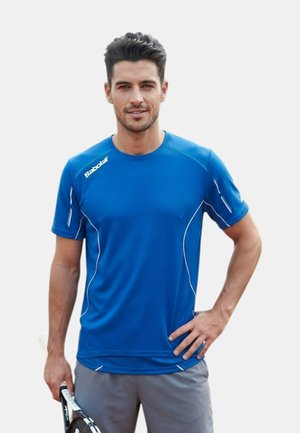 Uomo con maglia sportiva blu e pantaloncini grigi che tiene una racchetta da tennis con una mano sui fianchi, in piedi contro uno sfondo semplice.