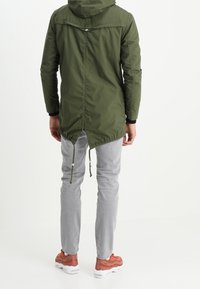 Oliwkowa zielona parka z kapturem, regulowanymi ściągaczami i dłuższym tylnym brzegiem. W zestawie z jasnoszarymi dżinsami i brązowymi butami sportowymi.
