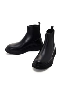 Une paire de bottes Chelsea noires en cuir avec des panneaux élastiques sur les côtés, des languettes de tirage et des semelles en caoutchouc épaisses sur fond blanc.