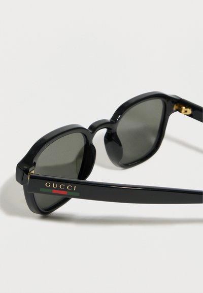 Lunettes de soleil géométriques noires avec verres foncés, arborant le logo "GUCCI" et une bande rouge-vert sur les branches, sur fond blanc.