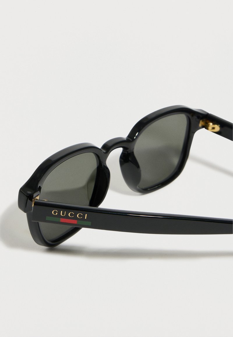 Lunettes de soleil géométriques noires avec verres foncés, arborant le logo "GUCCI" et une bande rouge-vert sur les branches, sur fond blanc.