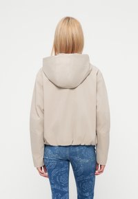 Μπεζ κοντό σακάκι με κουκούλα, με λεία, ματ υφή κα ι ελαστική μέση. Συνδυασμένο με μπλε τζιν με σχέδιο.