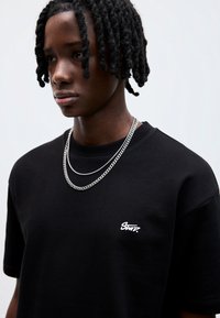 Sort bomulds T-shirt med korte ærmer og rund hals. Har et lille hvidt logo på brystet og lagdelte sølvkædehalskæder.