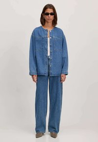 Jeansjacke und weit geschnittene Jeans in hellblau. Die Jacke hat einen runden Halsausschnitt und eine Knopfleiste mit zwei Brusttaschen. Sonnenbrille und High Heels.