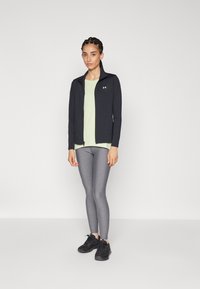 Chaqueta atlética negra con cremallera, top verde claro, mallas grises y zapatillas negras. Tejido suave, diseño ajustado y marca sutil en la chaqueta.