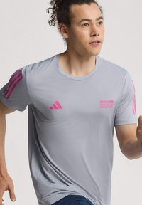 Muškarac s kovrčavom kosom vezanom unatrag nosi sivi BMW Berlin Marathon majicu s ružičastim Adidas logotipima, trči ili se jogira.