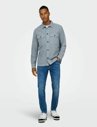 Camisa gris de botón con dos bolsillos en el pecho, llevada sobre una camiseta blanca, combinada con vaqueros azules y zapatillas negras y blancas.