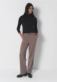 Donna in piedi contro un muro bianco, con indosso un top nero a maniche lunghe, pantaloni taupe a vita alta e larghi, scarpe nere, mani nelle tasche, sorridente.