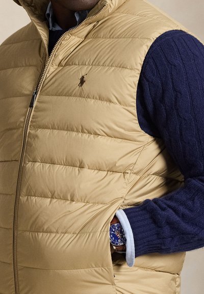 Bēšs pūkains veste ar horizontālu šuvumu, rāvējslēdzēja aizdari un izšūtu logotipu. Vilkts pāri tumši zilai kabatu džemperim ar gaiši zilu aproci.