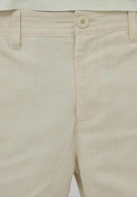 Pantalon en velours côtelé beige avec une fine texture côtelée verticale, doté d'une fermeture à un bouton et d'un détail de poche avant.