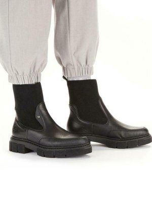 Black leather ankle boots with a chunky sole, featuring elastic side panels and a textured surface. Styled with light gray tapered pants.