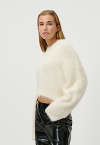 Pull court blanc et moelleux avec des manches longues, associé à un pantalon noir brillant. Texture douce et coupe décontractée.