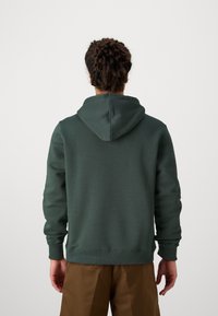 Dark green hoodie made of soft fabric, featuring a drawstring hood, ribbed cuffs, and hem; worn with light brown pants.