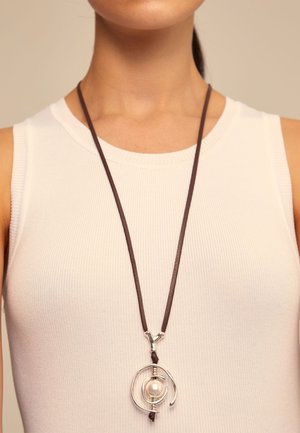 Mujer con top blanco sin mangas y un collar largo marrón oscuro con colgante de espiral plateada que sostiene una perla central.