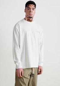 Weißes Langarm-T-Shirt mit Rundhalsausschnitt, versehen mit einem kleinen lila Logo auf der linken Brust. Der Stoff wirkt weich und leicht.