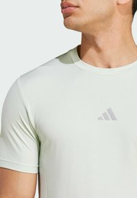 Světlově zelené sportovní tričko s krátkým rukávem, texturovaná látka a decentní šedé logo na hrudi. Detailní pohled na výstřih.