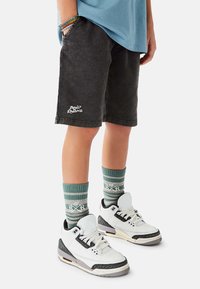 Mustat denimgolf-shortsit logoyksityiskohdilla, vihreä-valkoiset raidalliset sukat, valkoiset ja mustat lenkkarit harmailla yksityiskohdilla, litteä pohja.