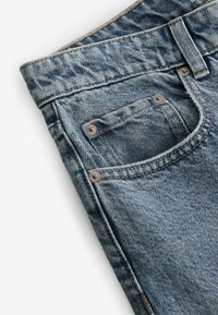 Jeans de denim en azul claro con una textura desvanecida. Las características incluyen un pequeño bolsillo con remaches de metal y costuras en contraste a lo largo de las costuras.