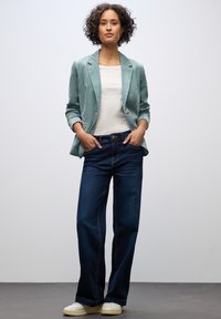 Vaaleanvihreä corduroy-takki valkoisen paidan päällä, yhdistetty tummansinisiin suoriin farkkuihin ja valkoisiin tennareihin. Yksinkertainen muotoilu, teksturoitu kangas.