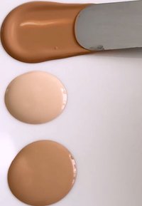 Kermainen nestemäinen meikkipohja kolmessa sävyssä: vaalean beige, keskiruskea ja syvän ruskea, levitetty Spatulan avulla sileälle valkoiselle pinnalle.