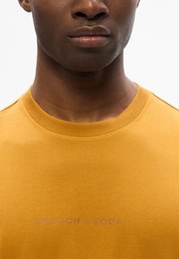 Sininen T-paita, jossa on pyöreä pääntie, valmistettu pehmeästä kankaasta. Edessä on painettu "SCOTCH & SODA" violetilla. Yksinkertainen muotoilu, lyhyet hihat.