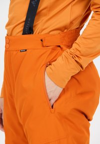 Des pantalons de ski orange en tissu résistant dotés d'une taille avec des boutons, de bretelles réglables et d'une poche latérale pour le rangement.