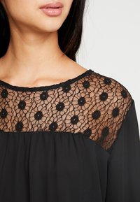Czarna bluzka z kwiatowym koronkowym karczkiem oraz gładkim, zwiewnym tułowiem. Koronka ma falisty brzeg i małe wzory kwiatowe.