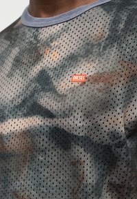 Άνδρας που φοράει γκρι και μαύρο πουκάμισο με διάτρητο δίχτυ και μικρό πορτοκαλί λογότυπο Diesel στο στήθος κοντά στον γιακά.