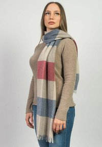 Ruteret tørklæde i beige, blå og røde nuancer. Lavet af blødt stof med frynsekanter. Båret draperet omkring halsen over en tan farvet sweater.