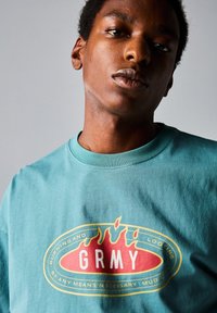 Camiseta de cuello redondo en color verde azulado con un estampado gráfico de llamas y el texto "GRMY" en rojo intenso, rodeado de un óvalo amarillo.