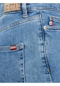 Detailní záběr na zadní kapsu modrých džínů z denimu značky Big Star, se značkovou visačkou s červenou hvězdou, prošitím a kovovým nýtem.