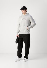 Siva dukserica s printom "HILFIGER NEW YORK" logotipa, crne hlače, bijele tenisice i crna kapa. Drži malu crnu torbu.