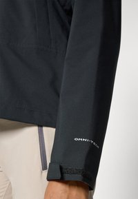 Black waterproof jacket sleeve featuring a smooth texture, adjustable Velcro cuff, and "OMNI-TECH" logo in white. Light-colored material underneath.