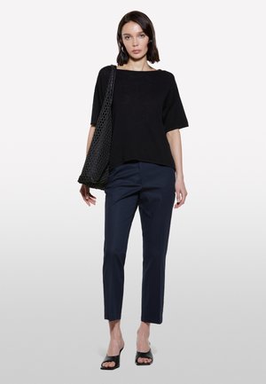 Top nero a maniche corte con scollatura a barca, abbinato a pantaloni blu scuro slim-fit. Il modello tiene una borsa a rete nera e indossa delle scarpe con apertura sulla punta nere.